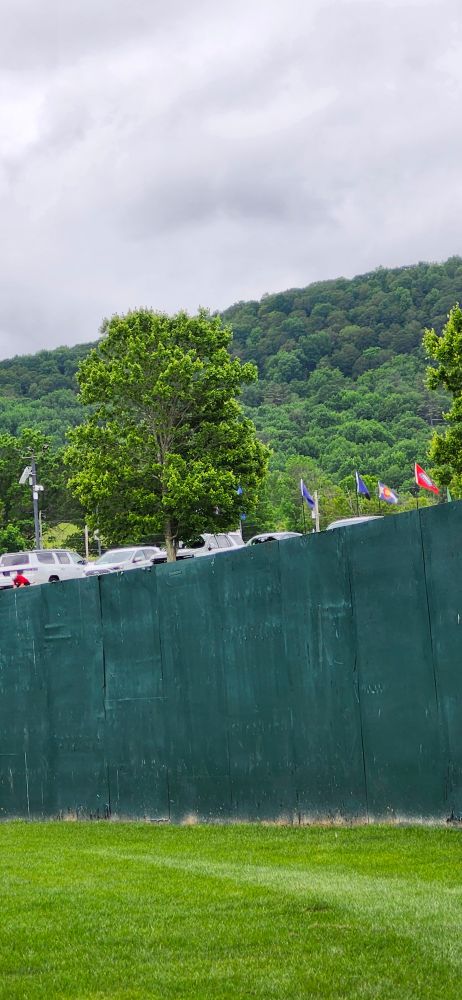 Gray skies, blue-green hills, and the outfield wall topped with flahs on a summer day. 