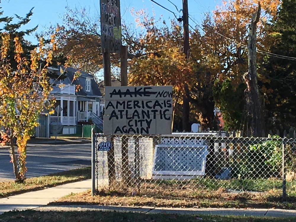 Photo of a sign on a chain link fence. Original handwritten text is “make America Atlantic City again”; the word make is crossed out and replaced with “now.”