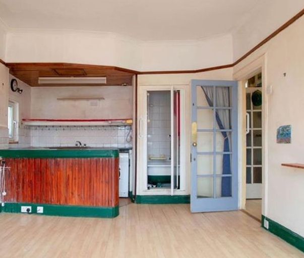 Estate agent photo of a living room / kitchen. In the corner next to the kitchen area is a doorway into a small room, inside which a bathroom is visible. There is a glass door into a hallway, and a glass entrance door to the flat beyond that.