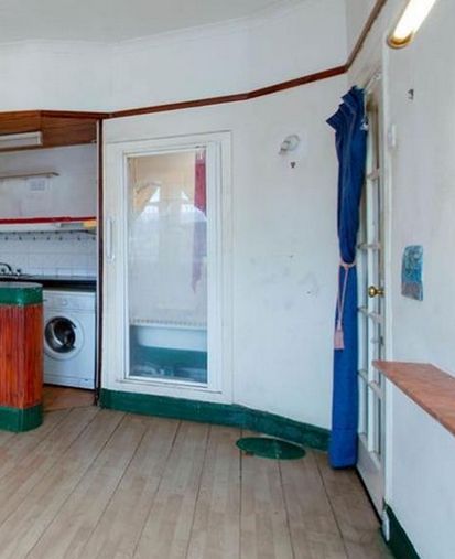 Estate agent photo of a living room / kitchen. In the corner next to the kitchen area is a glass shower door into a small room, inside which a bathroom is visible. There is a glass door into a hallway, and a glass entrance door to the flat beyond that.