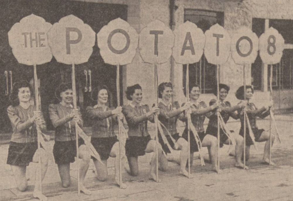 newsprint photo. 8 young women are kneeling down and hold up signs that spell out "The Potato Eight", each is in a matching gymanstic outfit. Behind them is an art deco swimming pavilion.