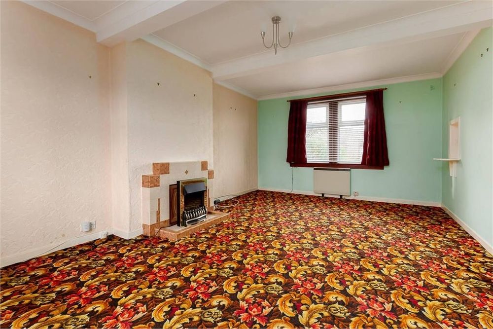 Estate agents photo of an empty sitting room. The walls are painted white and pale green. The carpet is a bold design of brown, yellow, orange and red with a pattern like feathers