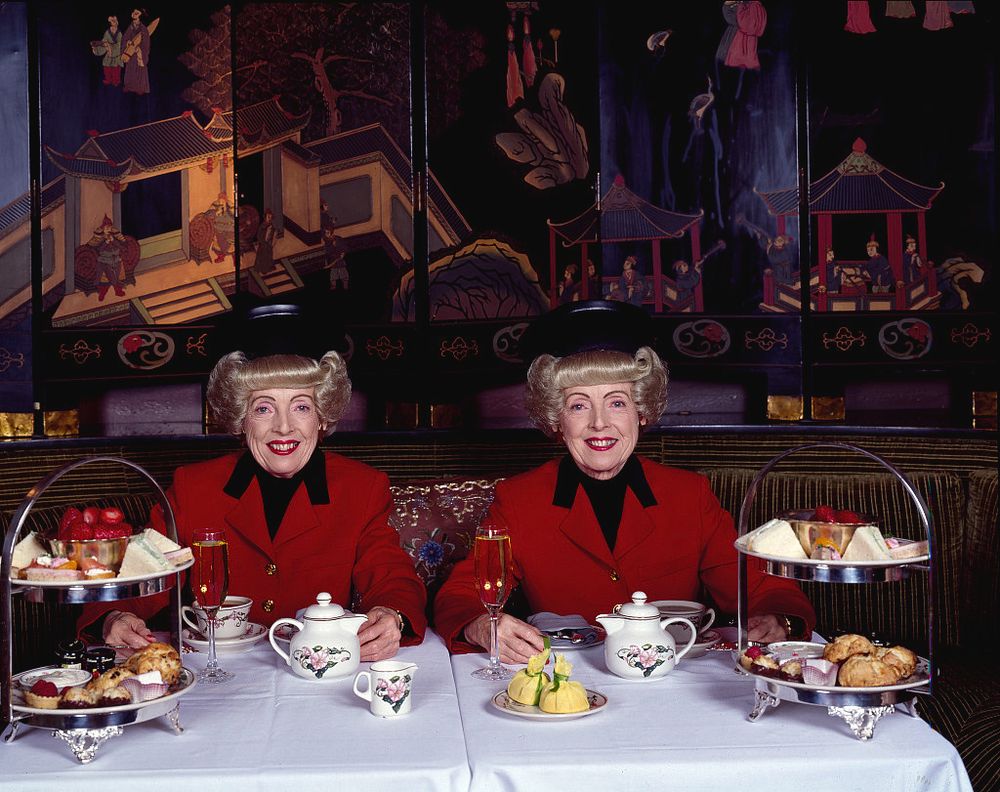 Twin older women wearing matching smart red jackets and elaborate blonde hairdos sit smiling at a table that is set up for afternoon tea.