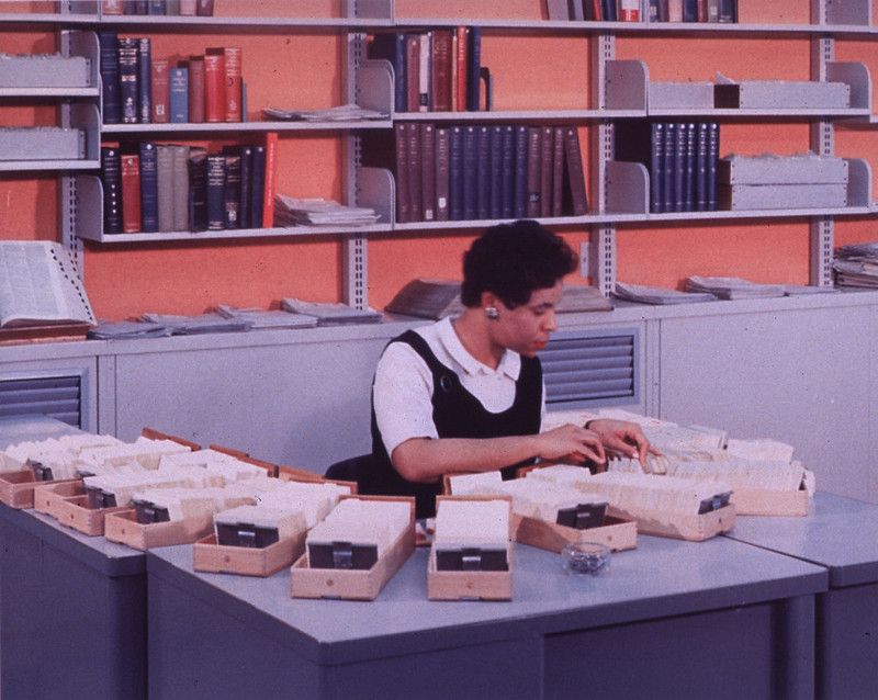 Interior view: Card catalog drawers are on the desk. Book shelves line the wall behind the desk. A woman in professional dress looks through a catalog drawer.