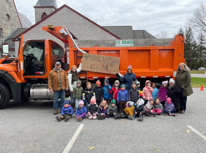 A group of school children sit in front of an orange truck which is a plow/sander. They are holding up a hand-drawn sign which I can't read but I think it says THANK YOU