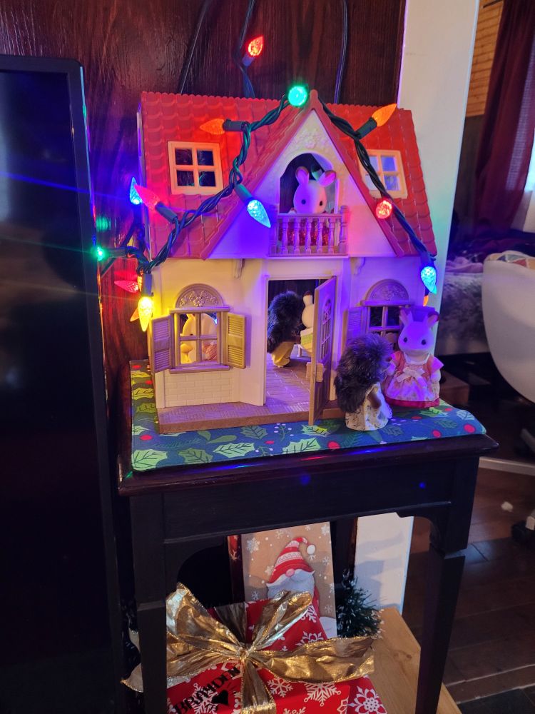 A close up of the calico critter cottage. A string of disproportionately large christmas lights wrap around the house's eaves and presents addressed to Brayden can be seen below the display. The cottage has several critters posed within. Porcupine and Bunny calico critter families sharing in their celebrations together.