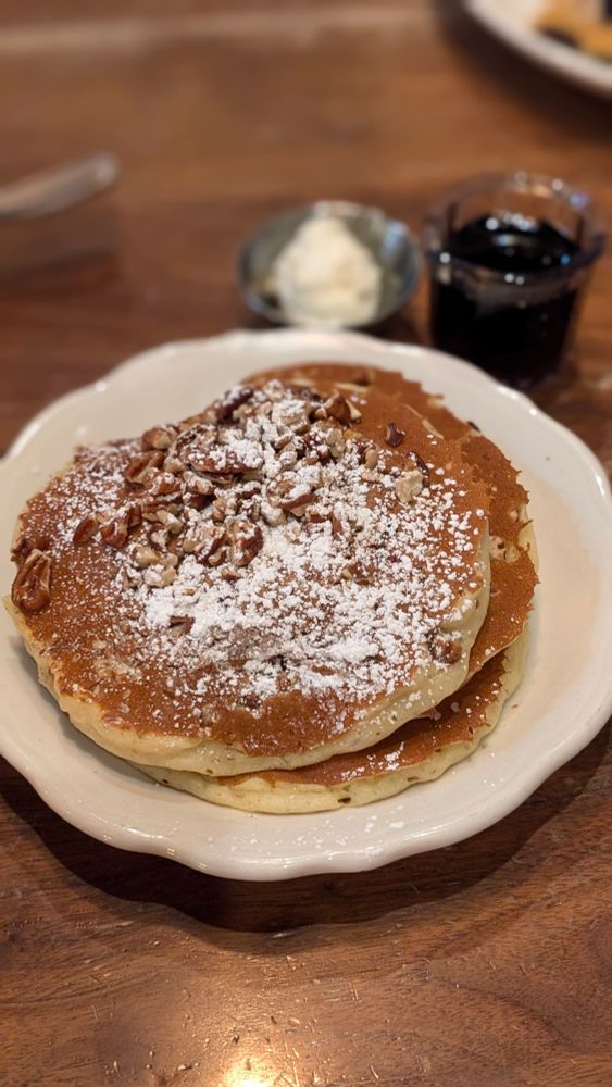 A glorious (if very slightly underdone) stack of three pecan pancakes on a white plate, topped with chopped pecans and a generous dusting of powdered sugar. Butter and syrup cups in the background, blurred to highlight the main event.