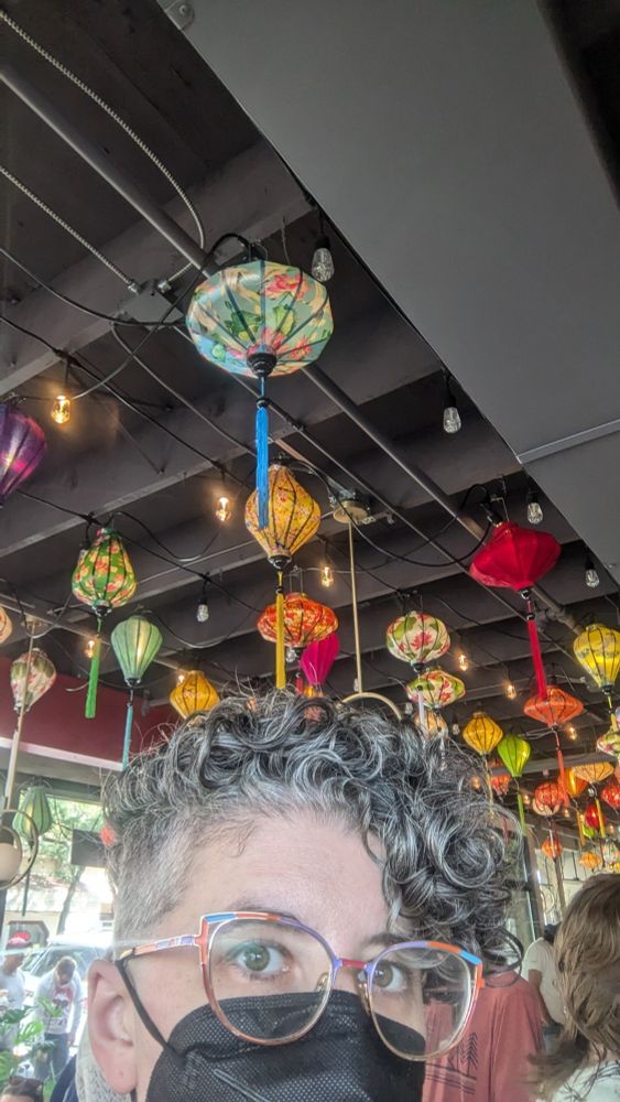 A curly-haired, spectacled person in a mask standing inside a hot, crowded cà phê place with pretty lanterns hanging from the ceiling
