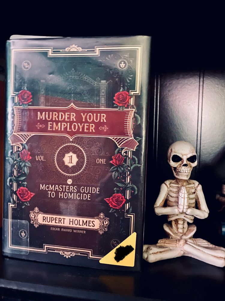 Book MURDER YOUR EMPLOYER by Rupert Holmes standing upright facing out on a black bookshelf next to a yoga skeleton figure.