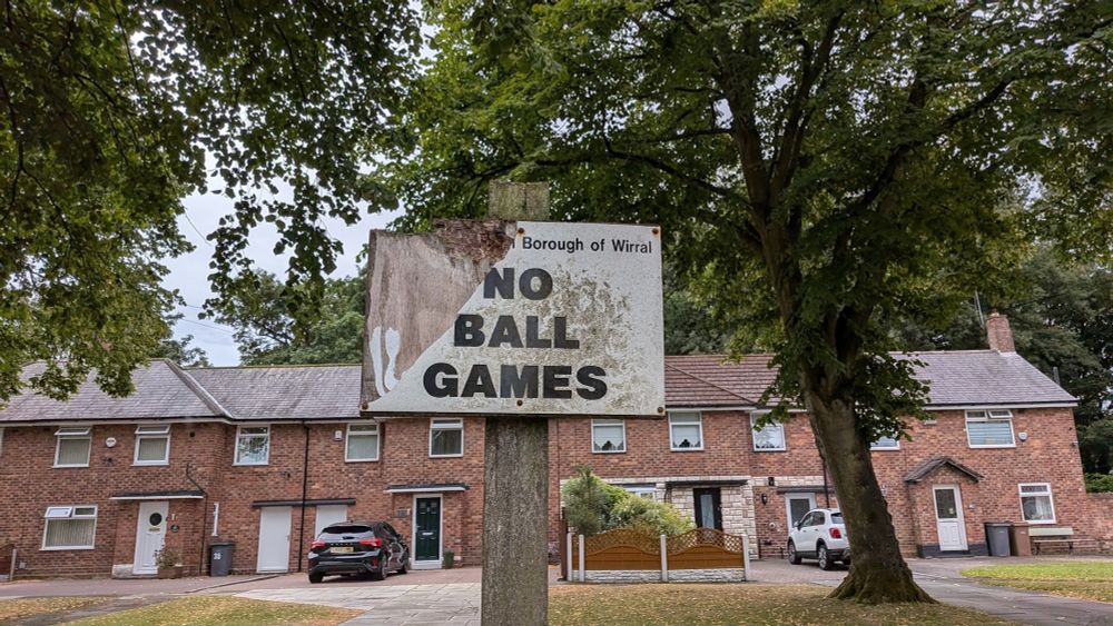 A broken sign that says 'NO BALL GAMES. Terraced houses and trees behind