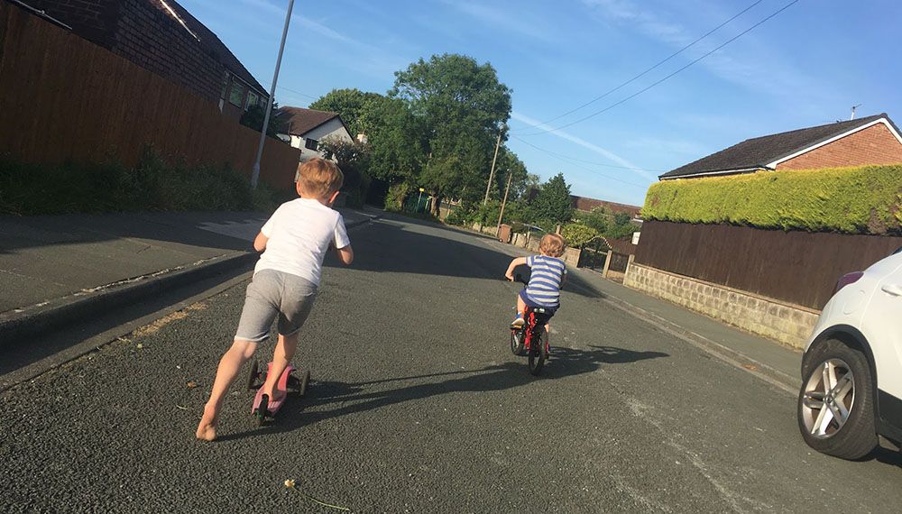 Two kids move along a quiet street in the sun. One on a scoot, one cycling