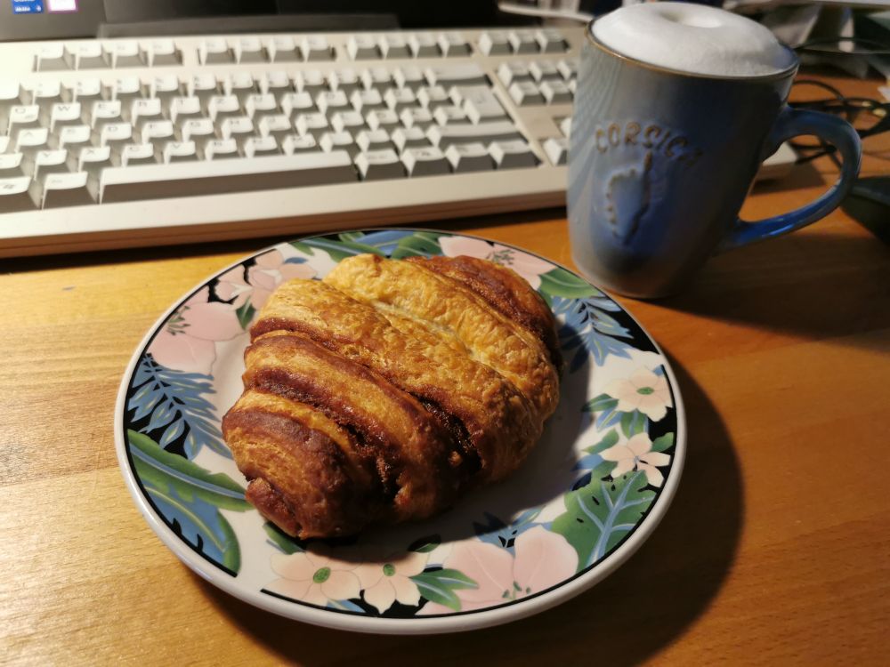 Auf einem Holztisch steht eine gefüllte hellblaue Korsikatasse mit Milchschaum und ein Teller mit einem Franzbrötchen.  Im Hintergrund eine hellgraue Computertastatur 