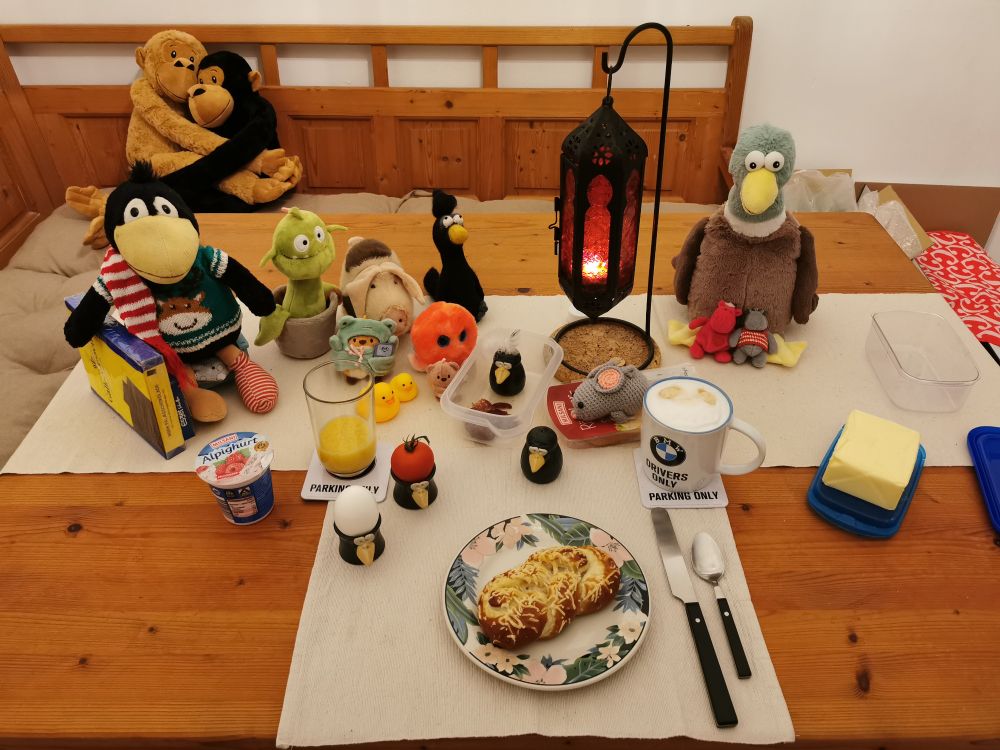 Ein gedeckter Frühstückstisch mit den beschriebenen Dingen, links sitzen ein Plüschrabe in grünem Strickpulli mit einem Rentierkopf darauf, der eine Packung blaugelbe Eszetschnitten hält und der eine rotweiß gestreiften Schal trägt, eine Monsterpflanze, ein Plüschschwein, ein Filzrabe, ein Plüschtentakel, ein Plüschfrosch, ein Plüschbärchen und zwei Gummienten. In Tischmitte steht eine rote, orientalische Kerzenleuchte, an deren Fuß eine hellgraue Häkelmaus auf dem Käse sitzt. Rechts der Lampe eine sehr große Plüschente, zwischen deren Füßen sitzen zwei Minihippos, ein rotes und ein graues mit rotweißem Pulli. Calimero (erster Rabeneierbecher) enthält das Ei, Kalimera (zweiter Rabeneierbecher) enthält eine Tomate. Munin (Rabenpfefferstreuer) bewacht die Salami, indem er im Behälter sitzt. Es gibt auch noch einen Rabensalzstreuer (Hugin). Die Butterschalen sind blau und grün. Auf der Eckbank hinter dem Tisch sitzen zwei sich umarmende Plüschaffen.