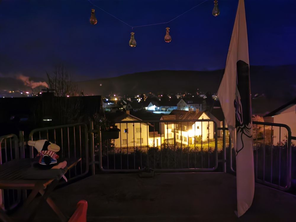 Blick über einen Balkon mit einem kleinen, bepflanzten grünen Blumenkasten auf dem Boden in der Mitte vor dem Geländer und einem dunkelgrünen bepflanzten Blumenkasten links am Geländer. Links steht eine rote Bank und ein Holztisch mit einem Plüschraben in grünem Strickpulli und rotweißem Schal mit wollweißer Mütze auf einem grauen Kissen sitzend darauf, rechts hängt eine große Korsikaflagge. Oben hängt eine Kordel mit vier Solarleuchten quer. Im Hintergrund ein Dorf in der Dunkelheit.