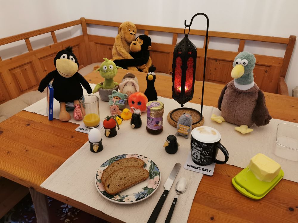 Ein gedeckter Frühstückstisch mit den beschriebenen Dingen, links sitzen ein Plüschrabe, der eine blaue Packung Eszetschnitten hält, eine Monsterpflanze, ein Plüschschwein, ein Filzrabe, ein Plüschtentakel, ein Plüschfrosch, ein Plüschbärchen und zwei Gummienten. In Tischmitte steht eine rote, orientalische Kerzenleuchte, an deren Fuß eine hellgraue Häkelmaus sitzt. Rechts der Lampe sitzt eine sehr große Plüschente. Munin (Rabenpfefferstreuer) zeigt uns beleidigt seine Rückseite. Ein Rabeneierbecher enthält eine Tomate, der andere ein Ei. Es gibt auch noch einen Rabensalzstreuer. Die Butterschale ist grün.