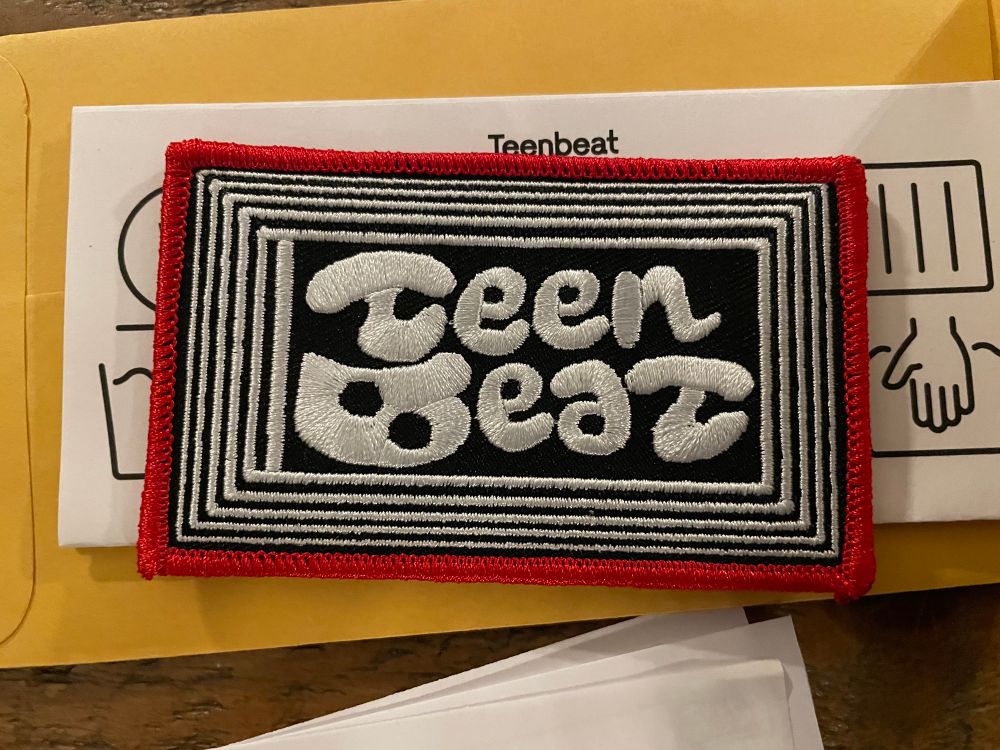 A patch with a black background and white lines around the center teenbeat logo. It is outlines in red and cool af. I need a denim jacket for this bad boy.