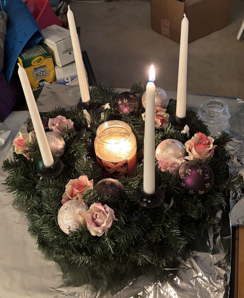 Advent wreath with green simulated pine, pink roses, and white Christmas ornaments. Around the ring are 4 white tapered candles, one of which is lit. In the center is a lit large jar candle.