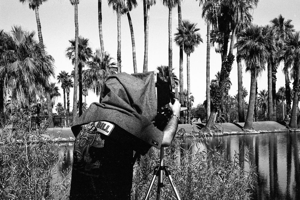A black and white photograph of a man working to focus a large format (8 x 10) camera. 
