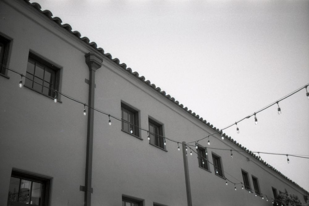 A black and white film photograph of a Spanish-style building. Pendant lights are strung across the courtyard that is out of frame. 