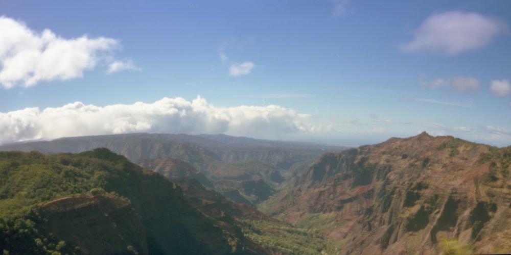 A panoramic film photograph taken on a wooden pinhole camera featuring Waimea Canyon on the island of Kauai. The green and red mountains fill the frame with a valley in the center. Large white clouds are scattered throughout the sky. 