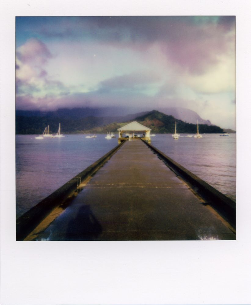 A Polaroid photograph of the Hanalei Pier. The mountains in the background a covered by mist and clouds from the morning rain. Purple hues are prominent in the ocean water. 