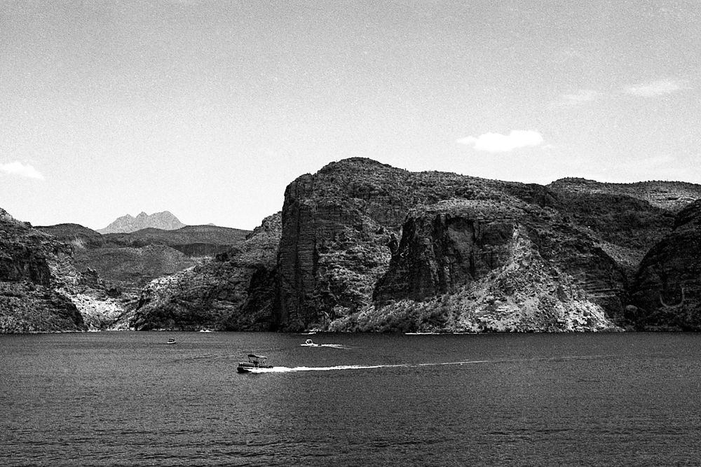 A black and white photograph of Canyon Lake in Arizona. Large rock formations surround the lake. A speedboat can be seen crossing the center of the frame.  