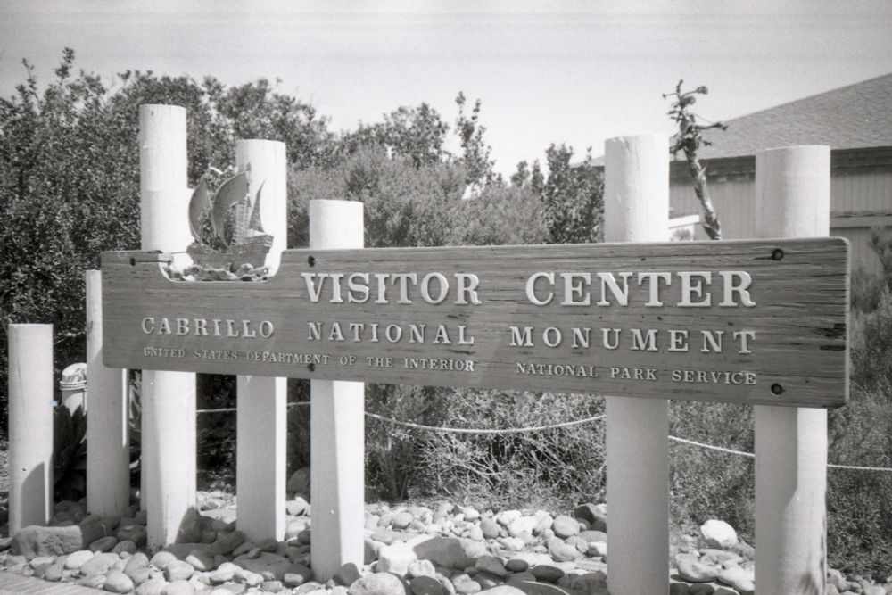 A black and white film photograph of the Visitor Center sign at Cabrillo National Monument in San Diego. The sign is wooden and mounted to several wooden posts. Shrubs and bushes grow behind it. 
