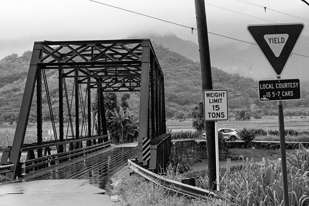 A black and white film photograph of the one lane bridge leading to Hanalei on the island of Kauai. It has a classic street truss structure. The wooden deck is glistening from the rain that just ended. Traffic signs can been seen that state “weight limit 15 tons” and “local courtesy is 5-7 cars”. 