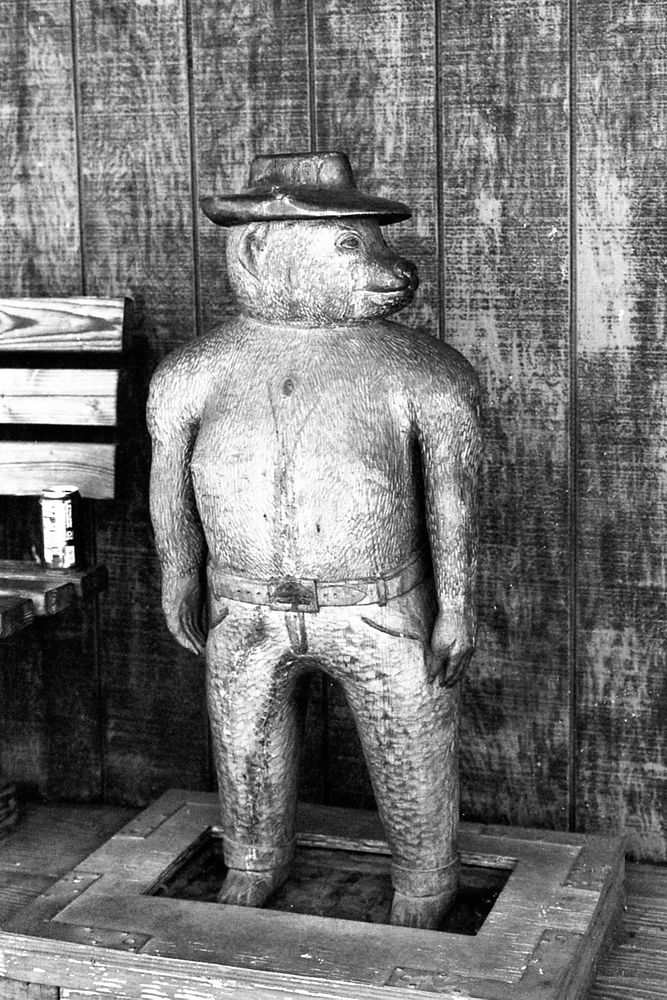 A black and white photograph of a wooden bear statue outside of the small stores in Tortilla Flat. He is about 3 feet tall with a hat, pants, and a belt and is looking right. 