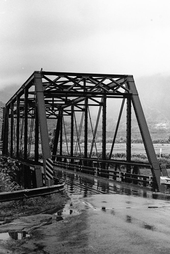 A black and white film photograph of the one lane bridge leading to Hanalei on the island of Kauai. It has a classic street truss structure with a wooden deck. The ground is glistening from the rain that just ended. 
