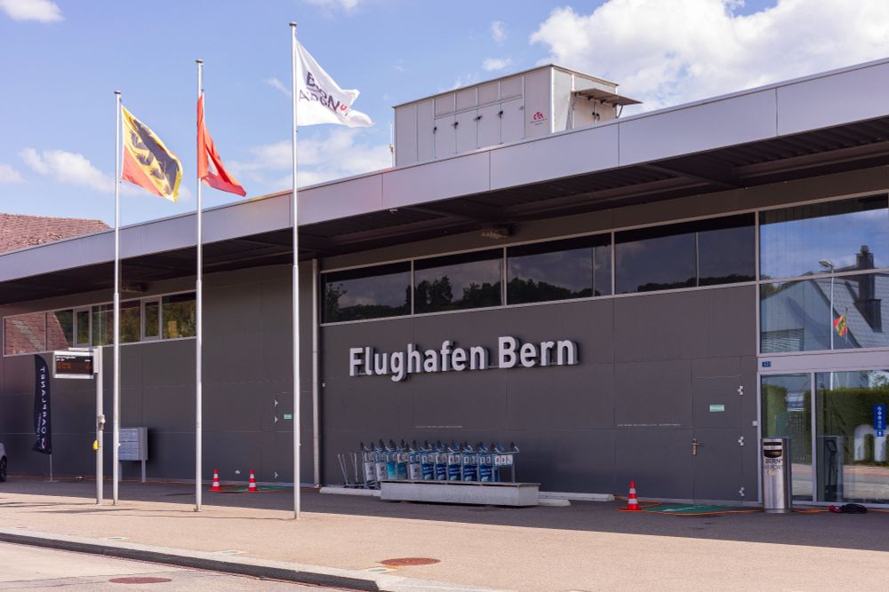 Aussenansicht des Terminals vom Flughafen Bern (Bern-Belp), aufgenommen am 5. August 2025. Zu sehen ist die moderne Fassade mit der Aufschrift "Flughafen Bern", davor mehrere Gepäckwagen und drei Fahnenmasten mit der Berner Kantonsflagge, der Schweizer Flagge und einer Flughafenflagge. Der Himmel ist leicht bewölkt mit sonnigen Abschnitten.

Exterior view of Bern Airport terminal (Bern-Belp), photographed on August 5, 2025, showing the modern facade, luggage carts, and three flags under a partly cloudy sky.