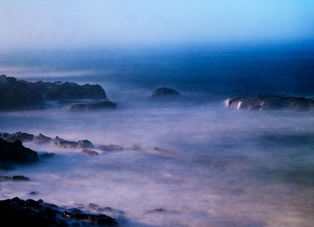 This serene coastal scene captures dark, rugged rocks surrounded by a dreamy, mist-like veil of water. The effect, achieved through a long exposure, softens the ocean into silky layers of pale lavender and blue. The gradient sky transitions from soft blue at the horizon to deeper blue above, enhancing the tranquil, ethereal mood of the morning seascape.
