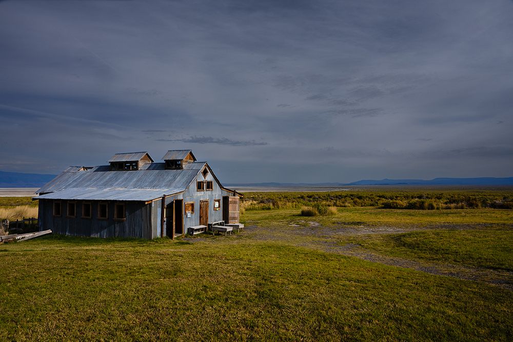 An old weathered building with corrugated metal walls and roof sits quietly in an open plain under a vast, moody sky. Golden light brushes the grass and distant hills, evoking a sense of solitude and timelessness in this remote landscape.