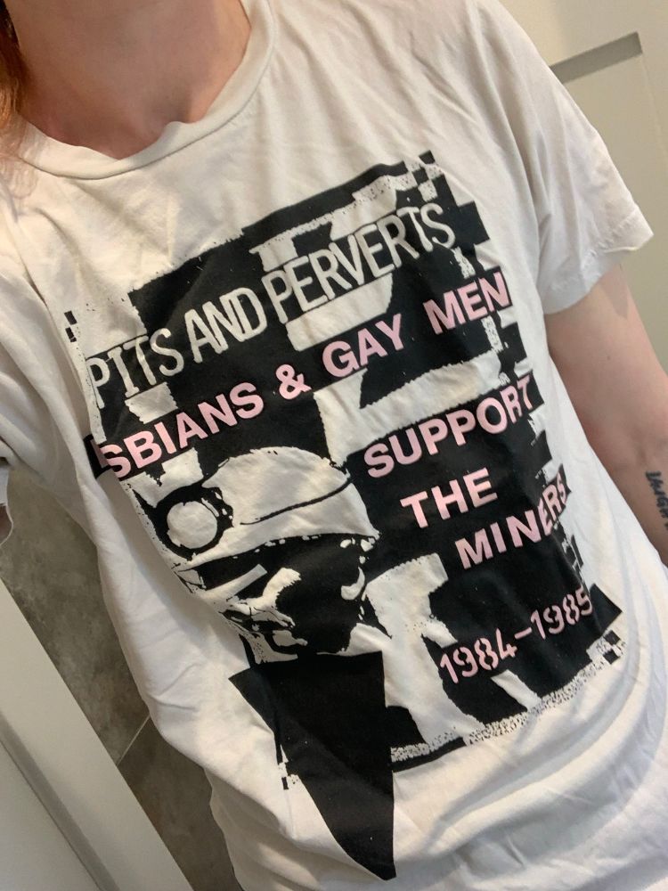 A white t-shirt with black and pink g graphics. The graphics show the head of a coal miner wearing a helmet along with an upside down triangle. Text reads “pits and perverts: lesbians and gay men support the miners. 1984-1985.”