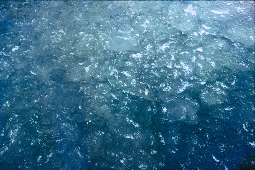 Fotografía analógica de la superficie del mar en invierno. El hielo que lo cubre aparece fragmentado en múltiples placas translúcidas, como un mosaico en tonos azulados y verdosos. Hay brillos dispersos donde la luz se refleja sobre las grietas y los cristales. La textura es granulada. 
