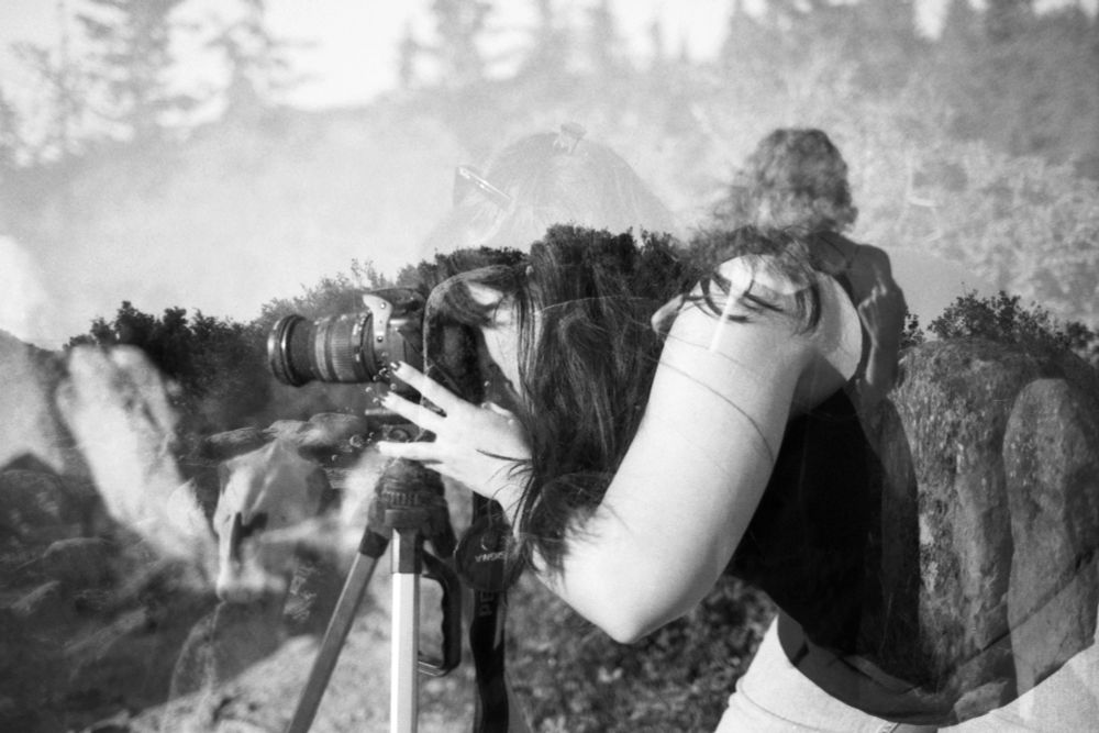 Black and white double exposure. Berta leans over a tripod-mounted camera, her silhouette blending with Iosune's dark figure, which looks like a shadow or a ghost. The backdrop showcases a wilderness scene with trees and boulders, adding depth to the ethereal, overlapping imagery.