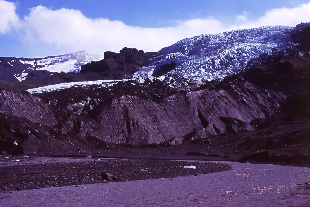 Another photograph of a glacier. The blue and white ice of the top layer gets blended with the brown body of the earth. It's a sunny day and the clouds embrace the glacier. The melted water that appears below is brown.