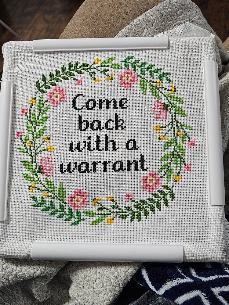 A white clip frame for needlepoint with a cross stitch design featuring a floral border and text that reads, "Come back with a warrant"
