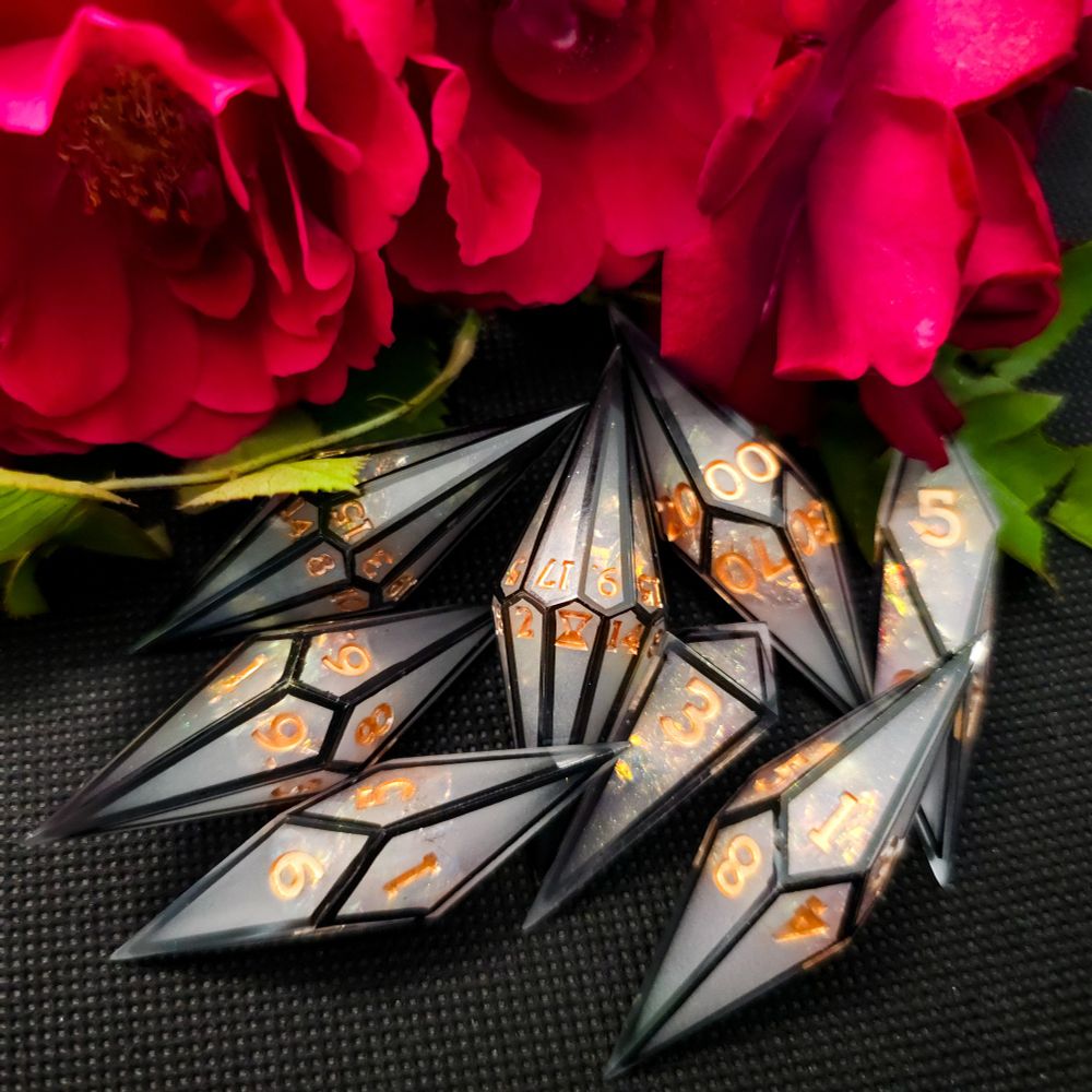 A set of crystal shaped polyhedral dice scattered in front of red roses. The dice are a foggy white color with black frames. The numbers are a bright brass color.