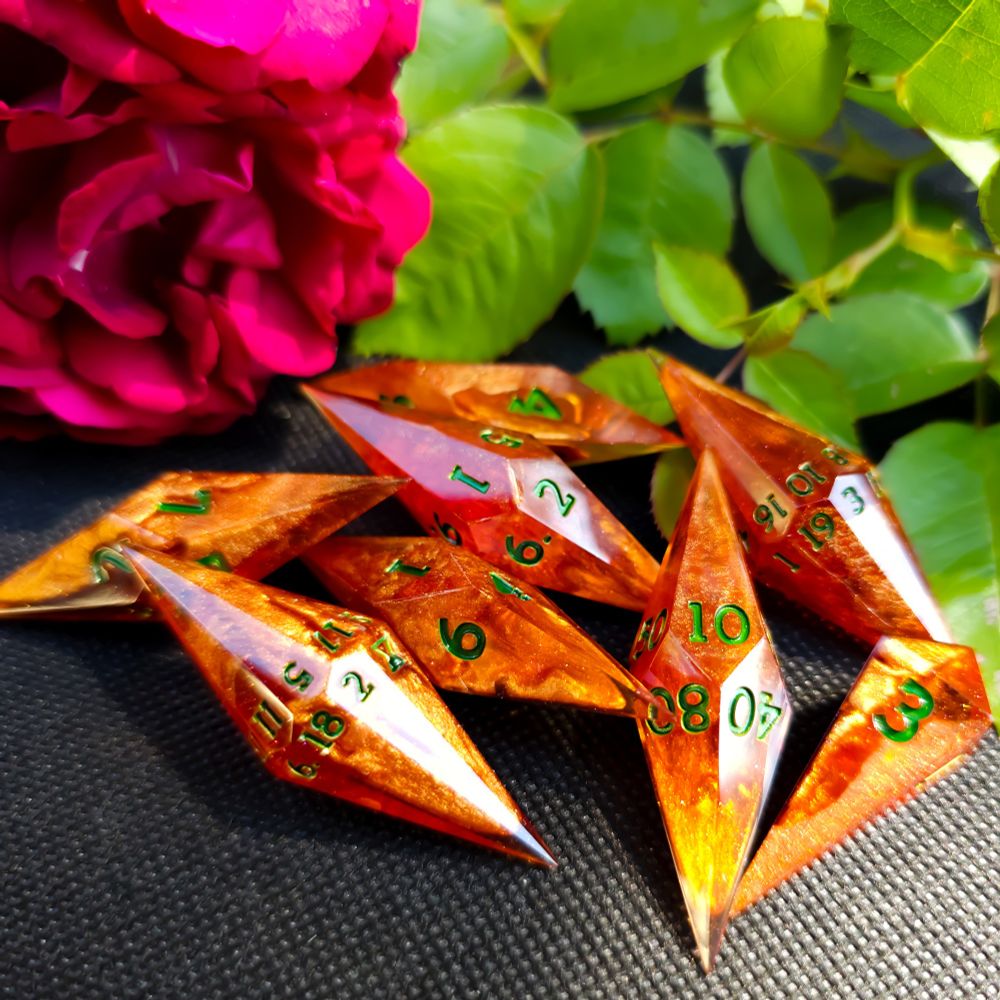 Vibrant orange crystal shaped dice with emerald green numbers lay on a black surface with red roses and greenery in the background.