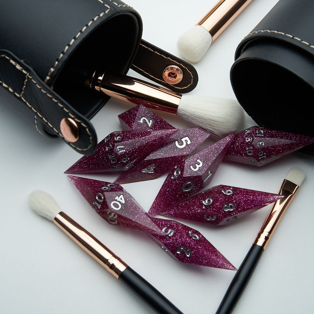 Crystal shaped dice made of dark pink glitter in resin with silver numbers lay scattered amidst makeup brushes and a black leather container on a white background 