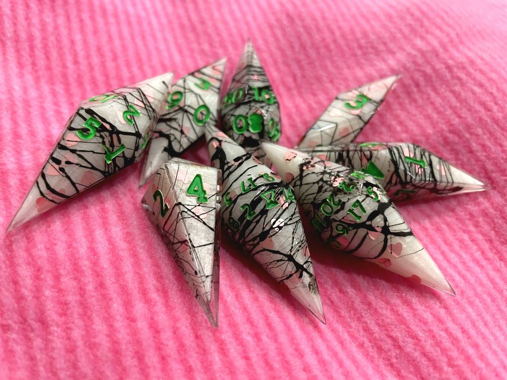 A set of pearlescent white crystal shaped dice with light pink hearts and chaotic wisps of black with bright green numbers on a striped pink background. 