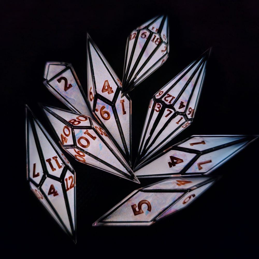 A set of glow-in-the-dark polyhedral dice scattered on a black background under no light. This set is glowing bright white, showing off the black frames and brass colored numbers. 