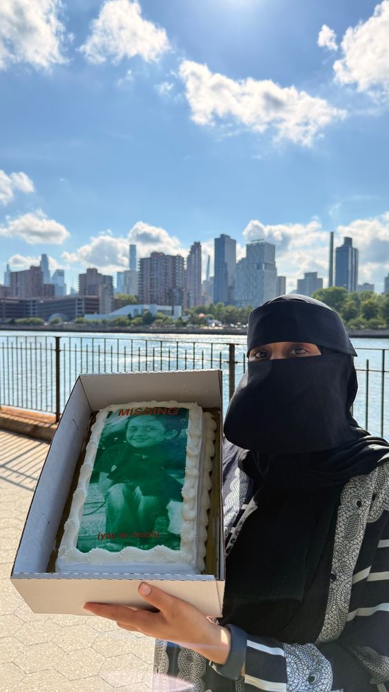 raheema standing in front of the city skyline holding a cake with a picture of young raheema that reads “missing…. you so much!”
