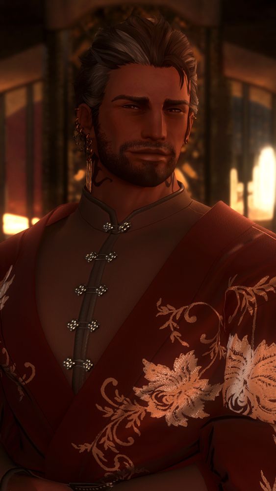 A screenshot of my oc, Krieg. He is a brown Highlander Hyur with brownish / amber eyes and dark brown hair with grey streaks. He has Archon marks on both sides of his neck.

He is dressed in a dark tan shirt that is clasped closed up the front, and leads to a semi high collar. Loose on his shoulders is a looser, dark red shirt that crosses in front of him, and is decorated with golden flower detailing.

He is smiling softly towards the camera as he stands with his arms clasped in front of him. The midday sun sneaks into the space through windows in the background