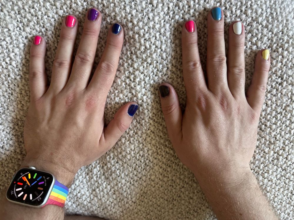 A photo of the author’s hands with painted fingernails. The left hand has pink, purple, and deep blue nails for bisexual pride, and also sports an Apple Watch with rainbow band and watch face. The right hand has deep purple, pink, light blue, white, and pale yellow nails for polyamorous pride. 