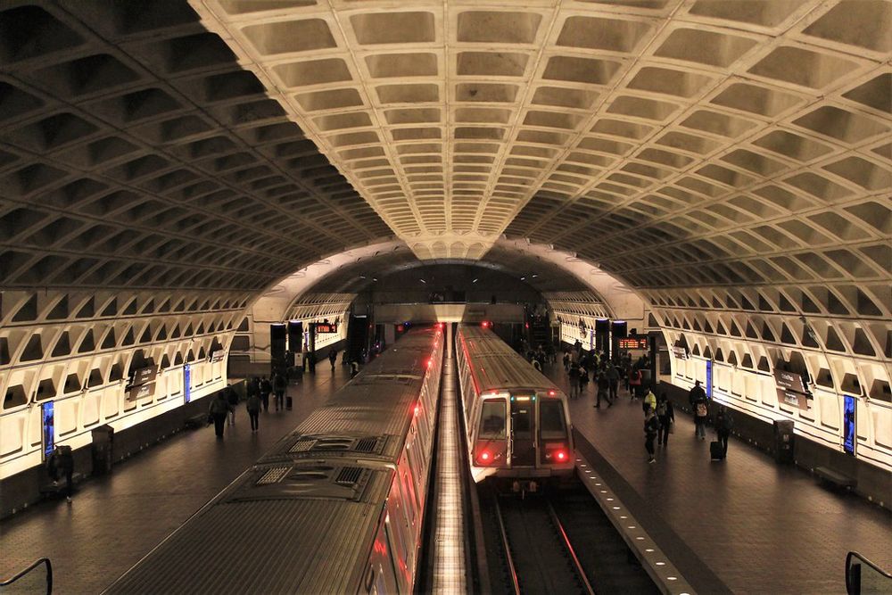 A photo of the interior of the L’Enfant Plaza Station of the WMATA subway in Washington, D.C., showing a coffered concrete ceiling, two trains at the platform extending forward, and openings for the perpendicular platforms hidden below the visible tracks. 