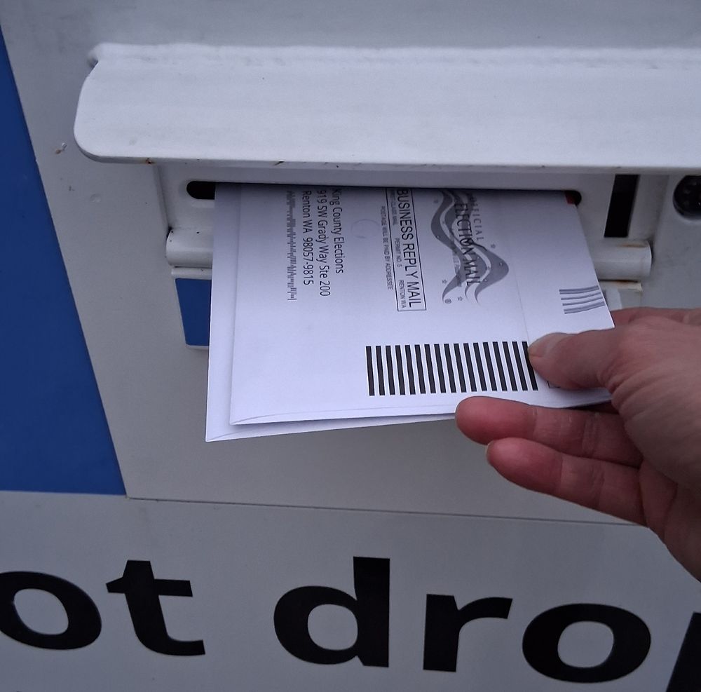A hand putting two ballots in a drop box. 