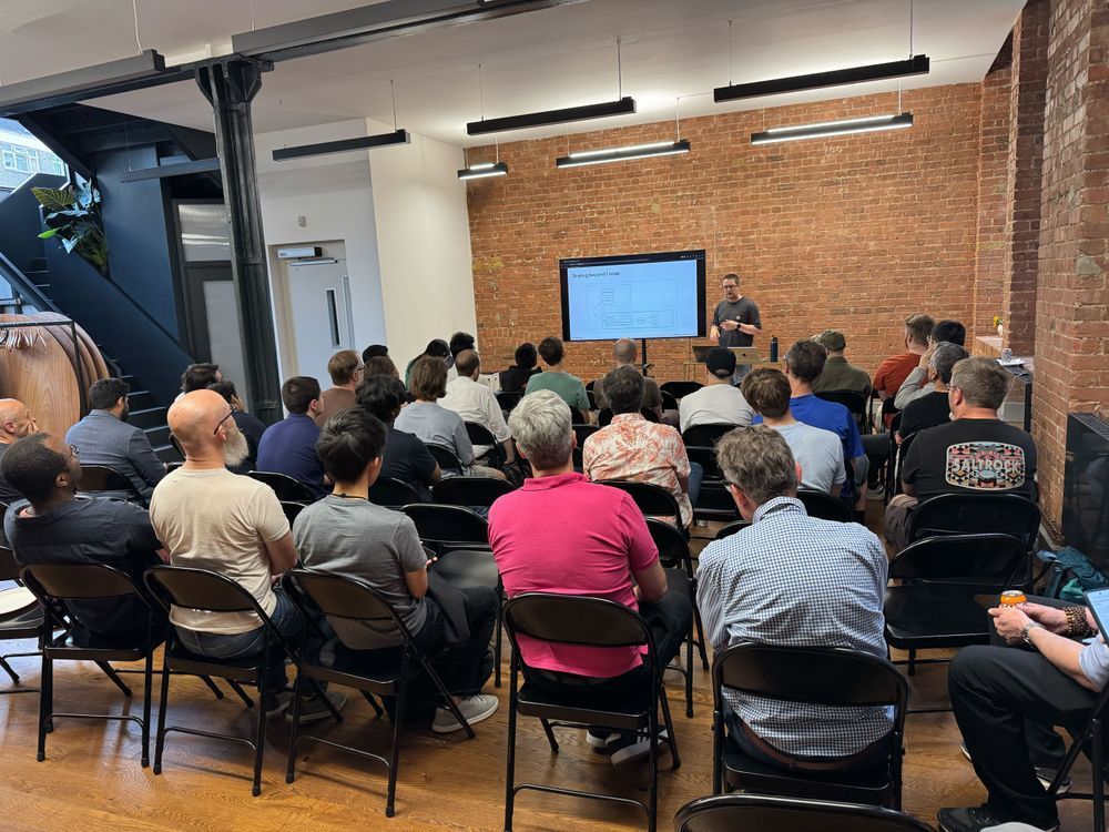 A speaker presenting to a full room at the Tailscale London Meetup, with attendees seated and focused on the talk, in a modern space with brick walls and industrial accents.