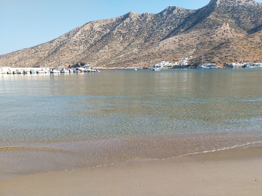 A view of a sea from the sandy beach and a rocky mountain in the background. On the left side of the horizon, there are some boats anchored at a small pier.  There is no wind, making it look like the sea is bleeding seamlessly with the sand. 